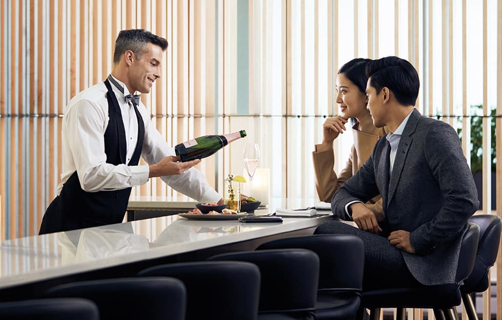 A server in an apron presents a bottle of champagne to two passengers seated at a table in Air Canada's Signature Suite, against a pale wood slat partition.