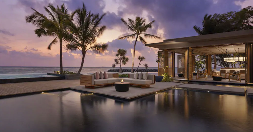 A luxurious outdoor lounge area by the ocean at sunset, featuring palm trees and elegant seating around a pool