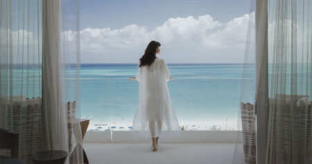 A woman standing on a balcony overlooking a beautiful ocean view, with sheer curtains framing the scene
