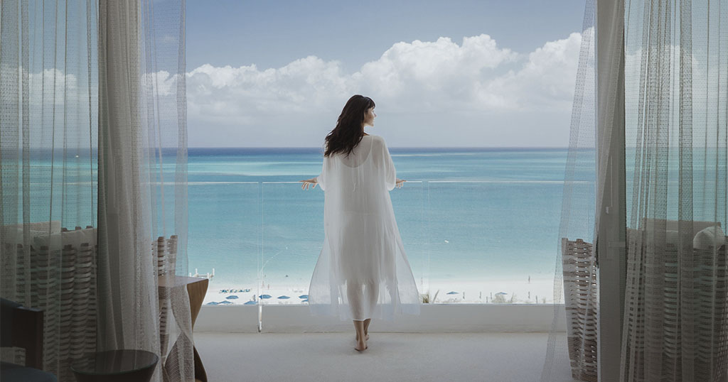 A woman standing on a balcony overlooking a beautiful ocean view, with sheer curtains framing the scene