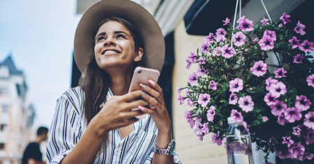 A smiling woman holding her phone, sitting outdoors near a pot of pink flowers