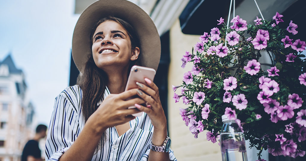 A smiling woman holding her phone, sitting outdoors near a pot of pink flowers