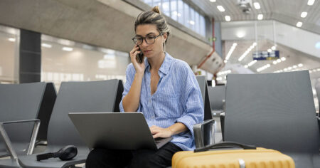 A woman sitting in an airport using a laptop and talking on her phone, with luggage nearby