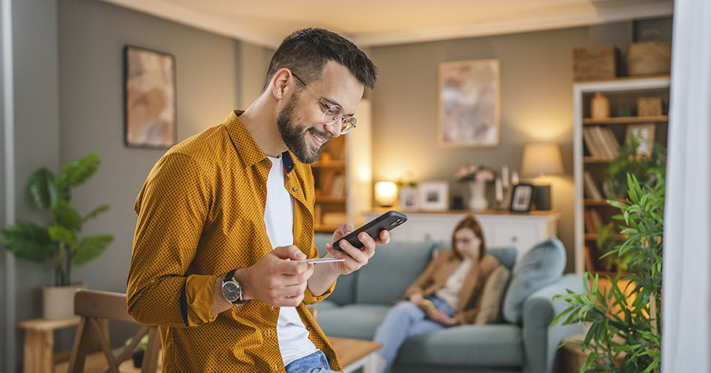 A man smiling while checking his phone, with a woman sitting on the couch in the background
