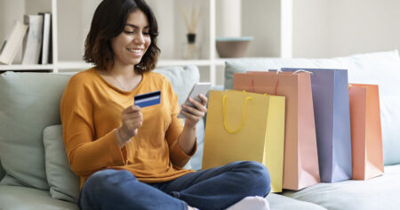 A person happily shopping online at home, holding a credit card and phone, with shopping bags beside them