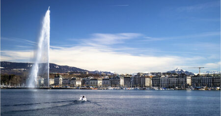 A large fountain sprays water high into the air, with a city and mountains in the background under a clear blue sky
