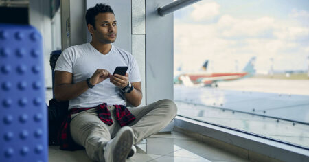 A man sitting on the floor at an airport, using his phone while waiting for a flight